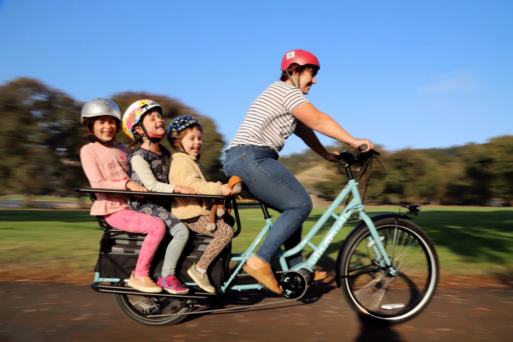 A view of a woman cycling the Xtracycle Swoop Electric Cargo Bike with chilren sitting having fun at the back