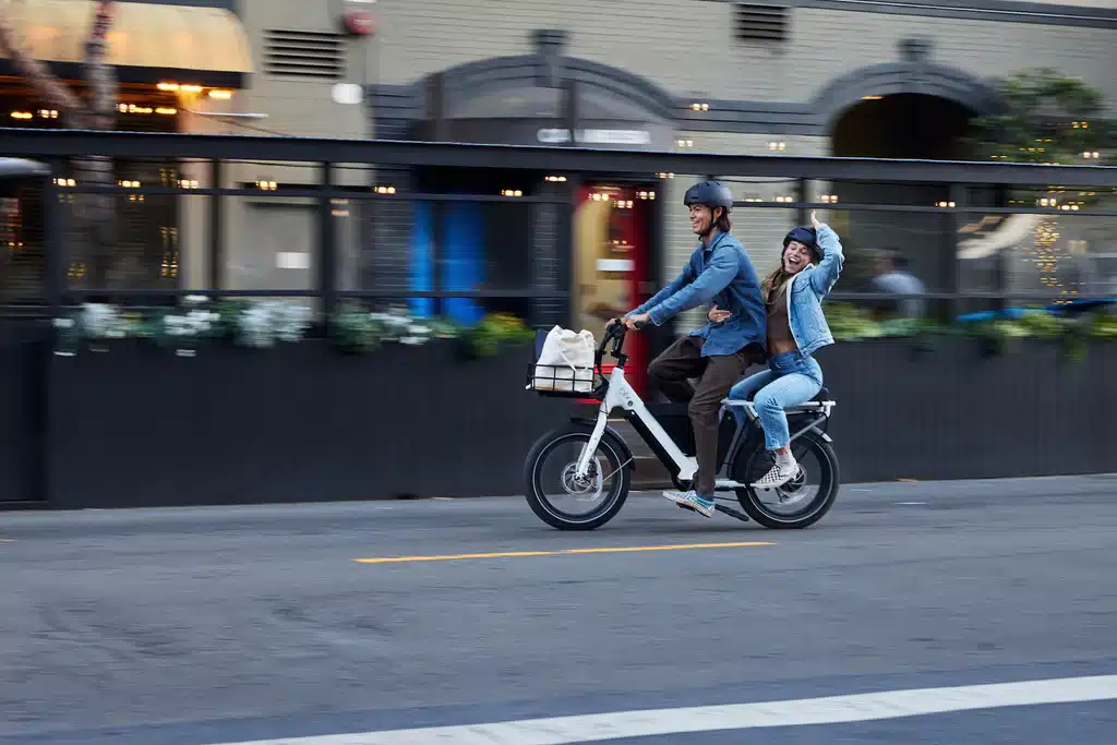 A mother teaching a kid how to ride an electric bike on a road with him sitting behind