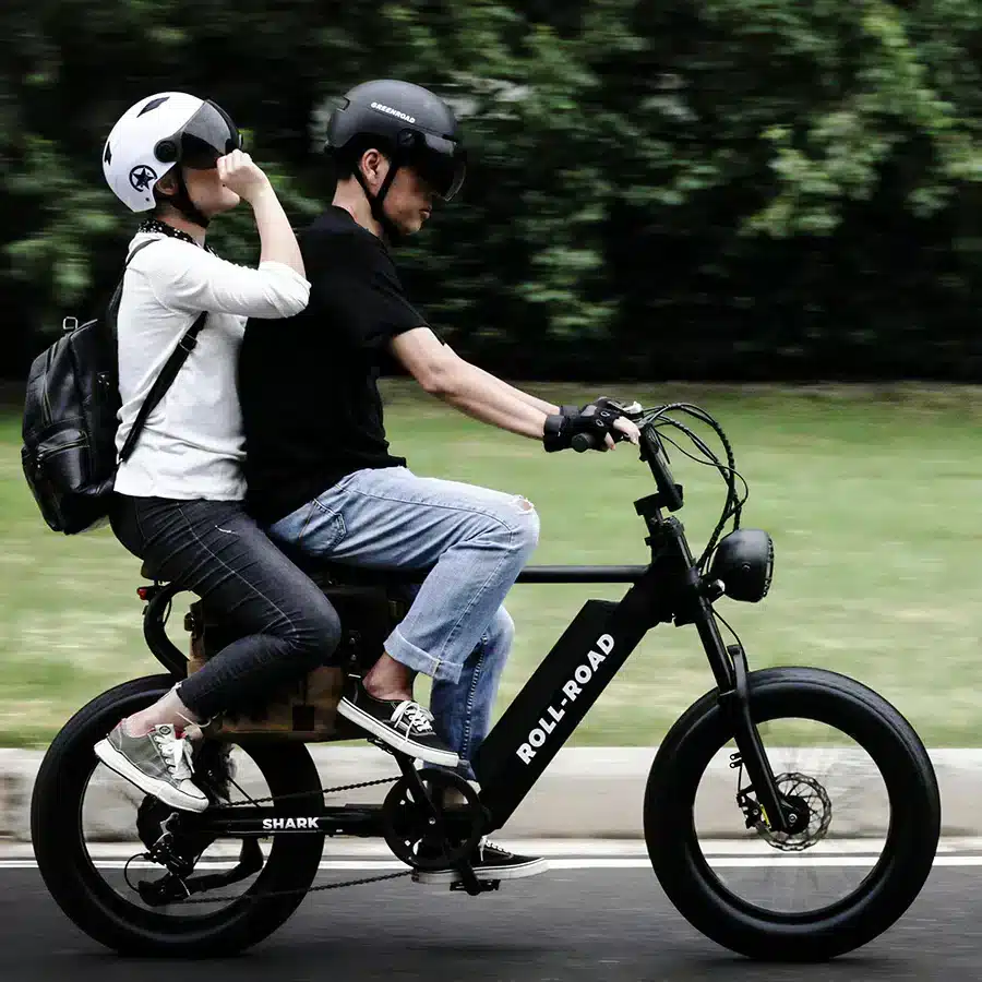 A side view of a couple riding a two seater bike wearing helmets